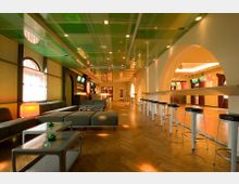 Moderne Lounge mit Holzfußboden, einer langen Sitzbank auf der linken Seite, einer Bar mit hohen Hockern rechts, und einer grünen Deckenkonstruktion. Große Rundbogenfenster und gewölbte Durchgänge verleihen dem Raum ein offenes Design.