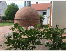 Ein runder, gemauerter Backsteinbau in einem gepflasterten Hof mit weißen Rosensträuchern im Vordergrund. Im Hintergrund eine grüne Wiese, ein Baum und helle Gebäude mit orangefarbenen Dächern.