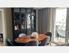 Essbereich in einer modernen Wohnung mit ovalem Holztisch und mehreren Polsterstühlen vor einem hohen schwarzen Vitrinenschrank. Rechts führen bodentiefe Glastüren mit grauen Vorhängen auf einen Balkon mit Metallgeländer und einem weißen Outdoor-Stuhl.