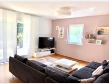 Ein modernes Wohnzimmer mit grauem Sofa, hellem Holzboden und einem weißen Couchtisch. Links befindet sich eine große Glastür mit dünnen Vorhängen, rechts ein Fenster und an der Wand ein Fernseher mit Regalen und Fotos.