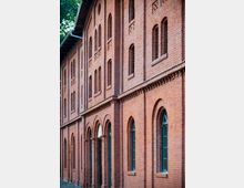 Backsteinfassade eines historischen Gebäudes mit hohen, schmalen Fenstern und Bögen. Die roten Ziegel und detaillierten Verzierungen wie ornamentale Mauerabschnitte prägen die Architektur.