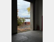 Blick aus einem schlichten, betonierten Innenraum durch eine große rechteckige Öffnung nach draußen auf ein Ufer mit herbstlichem Laub und Sträuchern. Dahinter liegt ein See mit einer gegenüberliegenden Stadt- oder Wohnbebauung unter bewölktem Himmel.