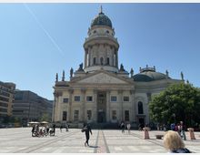 Barocke Kirche mit großer Kuppel und Statuen auf dem Dach, die sich auf einem gepflasterten Platz in einer städtischen Umgebung befindet. Im Vordergrund sind Fußgänger sowie einige Fahrräder zu sehen, dahinter moderne Gebäude und ein klarer, blauer Himmel.