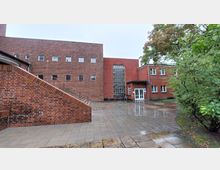 Backstein-Gebäudekomplex mit einer roten Fassade, rechteckigen Fenstern und einem großen Glasbaustein-Element in der Mitte. Davor ein gepflasterter, regennasser Hof mit Treppe und umgebenden Bäumen.