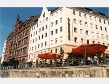 Straßenansicht mit historischer und moderner Architektur: Links ein rotes Gebäude im gotischen Stil, rechts ein heller, moderner Bau mit einem vertikalen Schild "Café". Im Vordergrund eine Terrasse mit roten Sonnenschirmen und Sitzgelegenheiten entlang eines Geländers, dahinter eine abschüssige Uferwand.