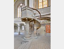Innenraum einer historischen Kirche mit einer verwitterten Kanzel, die über eine schmale, geschwungene Wendeltreppe erreichbar ist. Im Hintergrund sind hohe, bunte Glasfenster, dekorative Säulen und abgeblätterter Putz an den Wänden sichtbar.