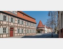 Straßenszene mit einem Fachwerkhaus im Vordergrund, gekennzeichnet durch hellrosa Fassaden, weiße Fensterläden und ein rotes Dach. Im Hintergrund ragt ein historisches Gebäude mit rotem konischen Dach hervor, umgeben von weiteren Altbauten und kahlen Bäumen unter klarem, blauem Himmel.