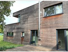 Moderne Holzfassade eines zweistöckigen Wohnhauses mit großen Fenstern und Schiebetüren, umgeben von einer grünen Wiese und Bäumen im Hintergrund. Eine Terrasse aus Holzdielen erstreckt sich vor dem Gebäude.