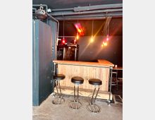 Eine kleine, minimalistische Bar mit einer Theke aus Holzlatten, drei schwarzen Barhockern mit Metallrahmen und mehreren hängenden Glühbirnen an der Decke. Im Hintergrund stehen Kühlschränke und Leuchtröhren sorgen für warme Beleuchtung.