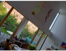 Großzügiger Wohnraum mit hohen Decken und großen bodentiefen Fenstern, die den Blick auf einen grünen Garten mit Bäumen gewähren. Im Innenraum befinden sich Möbel, Pflanzen, Dekorationen und ein Tisch, im Garten sind ein Holztisch und Stühle zu sehen.