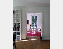 Wohnraum in einer modernen Wohnung mit weiß gestrichenen Wänden und Holzboden; im Vordergrund ein Bücherregal, im Hintergrund ein Essbereich mit rundem Tisch, vier weißen Stühlen, einem Blumenstrauß und einem Kronleuchter. Große Fenster lassen viel Tageslicht herein.