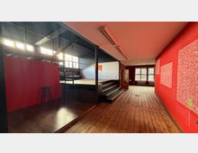 Innenraum einer ehemaligen Industriehalle mit breitem Holzfußboden, roten Wänden und einer erhöhten Plattform mit Treppe. Große Fensterreihen an der linken Seite und am Ende des Raums lassen viel Tageslicht hinein; dunkle Stahlträger und eine hohe Decke prägen die Architektur.