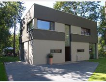 Zweistöckiges, modernes Wohnhaus mit rechteckiger Form, flachem Dach und einer Fassade in Beige und Dunkelgrau. Große Fenster und eine gepflasterte Einfahrt befinden sich vor dem Gebäude, umgeben von dichtem Baumbestand.