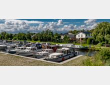 Kleiner Hafen mit mehreren festgemachten Motorbooten und Yachten, umgeben von Grünflächen und Häusern im Hintergrund. Der Himmel ist bewölkt mit sonnigen Abschnitten, und eine gepflasterte Uferpromenade führt entlang des Wassers.