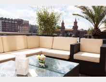 Moderne Outdoor-Sitzgruppe mit hellen Polstern und einem Glastisch, umgeben von Pflanzendekorationen wie Bambus und einer Palme, mit Blick auf die Oberbaumbrücke und die Spree in einem urbanen Stadtumfeld. Im Hintergrund sind historische Gebäude und der blaue Himmel sichtbar.