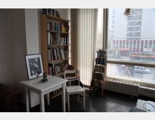 Ein kleiner, heller Raum mit einem Bücherregal, einem weißen Tisch mit Dekorationen und einem weißen Stuhl. Große Fenster mit vertikalen Jalousien bieten einen Blick auf eine Stadtstraße mit einem Gebäude im Hintergrund.