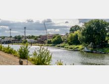 Industriegebiet am Flussufer mit Gebäudeanlagen, Kränen und Lagerflächen auf der einen Seite, sowie dichter Vegetation und kleinen Booten auf der anderen Seite des Wassers. Der Himmel ist leicht bewölkt, und die Flussoberfläche reflektiert das Licht der Sonne.