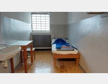 Kleiner, spartanisch eingerichteter Raum mit einer schmalen Liege mit blauer Decke, einem Holztisch und Hocker sowie einem Waschbecken. Ein vergittertes Fenster lässt gedämpftes Licht in den Raum.