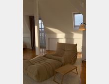 Ein minimalistisches, helles Wohnzimmer mit schräger Decke, einem beige gepolsterten Relaxsessel, einem passenden Fußhocker und einem kleinen Holztisch. Tageslicht fällt durch ein Dachfenster und eine verglaste Balkontür, die von einem braunen Vorhang umrahmt ist.