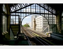 Blick von einer historischen Bahnstation auf erhöhte Gleise, mit einem Bogendach und Glasfenstern im Vordergrund. Im Hintergrund sind moderne Gebäude und verzierte Pfeiler entlang der Schienen zu sehen.