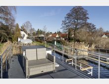 Hausboot an einem Wasserweg, umgeben von Schilf und einem Steg, im Hintergrund eine Siedlung mit Einfamilienhäusern und Gärten sowie Bäumen. Auf dem Deck steht eine graue Rattan-Sitzbank.