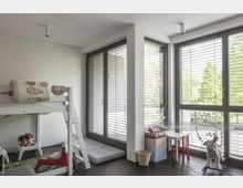 Kinderzimmer mit Hochbett und Spielbereich. Helle Glastüren mit grauen Rahmen und Jalousien führen zu einem Außenbereich; der Raum ist mit Spielsachen, einem kleinen Tisch mit Stuhl und einer Schaukelpferdfigur ausgestattet.