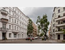 Straßenszene mit historischen Wohngebäuden im Altbau-Stil, verziert mit Stuckfassaden und Balkonen, flankiert von Bürgersteigen mit Bäumen. Im Hintergrund stehen parkende Autos, und ein bewölkter Himmel vervollständigt das städtische Bild.