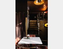 Modern eingerichtetes Café oder Restaurant mit geringer Beleuchtung. Im Vordergrund steht ein weißer Tisch mit roten Stühlen, dahinter ist eine Treppe zu einem oberen Bereich sichtbar, umgeben von dunklen Vorhängen und warmem, indirektem Licht.