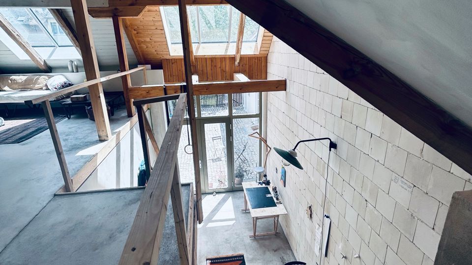 Blick in einen hohen, loftartigen Innenraum mit sichtbaren Holzbalken, geneigter Decke und großem Dachfenster sowie bodentiefen Glasfronten am Ende. Eine schmale Galerie mit Holzgeländer führt entlang der Wand; unten stehen wenige Möbel auf Betonboden vor einer hellen Ziegelwand.