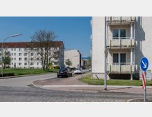 Straßenszene in einem Wohngebiet mit mehrstöckigen Wohngebäuden, verputzten Fassaden und Balkonen; links eine Straße mit parkenden Autos, rechts ein Rasenstreifen und ein Verkehrsschild. Im Hintergrund sind weitere Gebäude und ein Baum zu sehen.