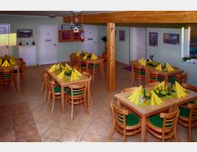 Ein kleiner Speiseraum mit Holztischen und passenden Stühlen, dekoriert mit gelben Servietten und Blumenarrangements. Die Wände sind hellgrün gestrichen, es hängen Gemälde und Dekorationen, und auf der rechten Seite befindet sich ein Fenster mit Tageslicht.