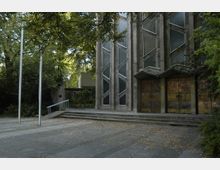 Modernes Kirchengebäude mit geometrischen Glasfenstern und kunstvoll verzierten Holztüren, umgeben von Bäumen und einem gepflasterten Vorplatz mit leichten Treppenstufen. Links befinden sich zwei hohe Fahnenmasten und ein Geländer.