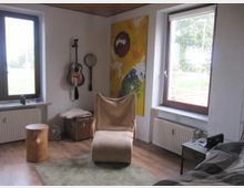 Wohnzimmer mit Holzboden und einer beige gepolsterten Liege. An der Wand hängen eine Gitarre und ein Banjo, daneben ein abstraktes buntes Gemälde; die Fenster lassen viel Tageslicht herein, mit Blick auf eine grüne Außenlandschaft.
