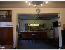 Eine Kneipe mit einer einfachen Bar im Hintergrund, beleuchtet von einem Schild mit der Aufschrift „Kindl Pils“. Es gibt Holzmöbel, wenige Barhocker, kleine runde Tische und Wanddekorationen wie ein Landschaftsgemälde und eine Uhr.