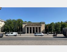 Ein neoklassizistisches Gebäude mit Säulenvorhalle und dreieckigem Giebel, umgeben von Bäumen. Im Vordergrund verläuft eine Straße mit parkenden und fahrenden Fahrzeugen, und einige Personen stehen vor dem Eingang des Gebäudes.