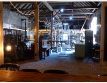 Ein rustikales, offenes Gebäude mit Holzbalken, unverputzten Ziegelwänden und einer hohen Decke. Im Raum stehen antike Möbel, ein Klavier, alte Maschinen und Beleuchtungskörper; Lichterketten und ein Dekobaum verleihen der Szene Gemütlichkeit.