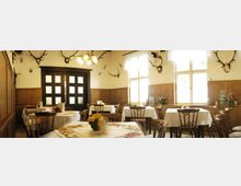 Ein traditioneller Gastraum mit Holztischen und -stühlen, dekoriert mit weißen Tischdecken und Blumenvasen. An den Wänden hängen Geweih-Trophäen, und große Fenster sowie eine Holztür im Hintergrund lassen Tageslicht herein.