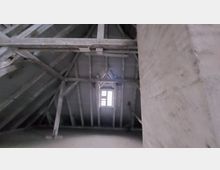 Dachboden mit sichtbarer Holzbalkenkonstruktion und hellem Fenster in der Mitte, das schwaches Tageslicht hereinlässt. Die Wände und das Dach wirken roh und unbehandelt.