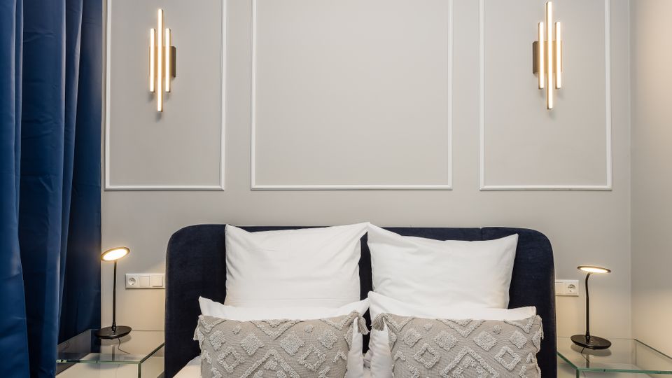 Modernes Schlafzimmer mit dunkel gepolstertem Bett, weißen Kissen und gemusterten Zierkissen vor einer hellgrauen Wand mit Zierleisten und zwei vertikalen Wandleuchten. Links hängen dunkelblaue Vorhänge; neben dem Bett stehen Glas-Nachttische mit schlanken Leselampen.