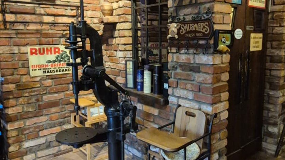 Rustikaler Innenraum mit freiliegenden Backsteinwänden, Ziegelboden und Holzdecke, mit einem gemauerten Rundbogen über einer Tür. Im Vordergrund steht eine große schwarze Handhebelpresse, daneben einfache Stühle; Regale und Wandflächen sind mit Flaschen und Vintage-Schildern dekoriert.