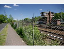 Gepflasterter Gehweg neben einer Straße, die von grünem Gras und kleinen Bäumen gesäumt ist. Rechts vom Gehweg ist ein Maschendrahtzaun, der ein Bahngelände mit Gleisen und einem Backsteingebäude umfasst, vermutlich ein Stellwerk.