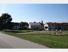 Kleiner Park mit Gehwegen, Grasflächen und einer kreisförmigen Blumenbepflanzung im Vordergrund; im Hintergrund sind Wohnhäuser mit roten Ziegeldächern, ein Kirchturm und Bäume zu sehen. Ein gepflasterter Weg verläuft am linken Bildrand.