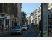 Straßenszene in einer Stadt mit mehrstöckigen Wohn- und Geschäftshäusern, einem Gehweg mit Fußgängern und Geschäften wie „Schlecker“ im Vordergrund. Auf der Straße fahren Autos und eine Straßenbahn, daneben steht ein Haltestellenschild der öffentlichen Verkehrsmittel.