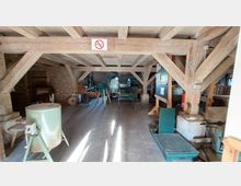 Innenansicht einer historischen Werkstatt mit Holzbalkenkonstruktion. Zu sehen sind verschiedene Maschinen und Geräte, darunter Metall- und Holzausstattung, auf einem Betonboden; an der Decke ein Schild mit Rauchverbotssymbol.