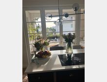 Moderne Küche mit einer weißen Kücheninsel, auf der ein Induktionskochfeld, ein Obstteller und zwei Blumenvasen stehen. Im Hintergrund sind große Fenster mit Blick auf eine sonnige Terrasse und ein Wohnviertel mit weißen Gebäuden zu sehen.