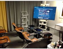 Ein kleiner Konferenz- oder Schulungsraum mit mehreren Ledersesseln, einem großen Flachbildschirm auf einem Rollständer, grauen und gelben Vorhängen im Hintergrund sowie kleinen Glasbeistelltischen. Im Raum stehen außerdem ein weißer Tisch, Lautsprecher und diverses technisches Zubehör.