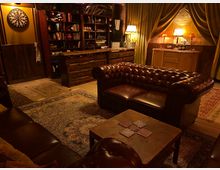 Gemütlicher Raum im Vintage-Stil mit braunen Chesterfield-Sofas, einem Holztisch und Teppichen auf Holzboden. Im Hintergrund eine rustikale Holztheke, ein Bücherregal, Vorhänge und eine beleuchtete Kommode an einer Backsteinwand.