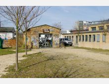 Ein kleiner Backsteinbau mit einer Glasfront und der Aufschrift „fabrik strand“; die Fassade ist stark mit Graffiti bedeckt. Im Vordergrund befindet sich eine sandige Fläche mit vereinzelten Bäumen, während im Hintergrund Bürogebäude und ein geparkter Van zu sehen sind.
