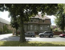 Straßenansicht eines historischen Wohnhauses mit dunkler Fassade, roten Dachziegeln und einem halbrunden Erker. Im Vordergrund stehen geparkte Autos und große Bäume, die Teile des Hauses beschatten; daneben ist ein mit Efeu bewachsener Backsteinbau zu sehen.