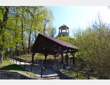 Ein steinerner Weg mit Geländer führt zu einem überdachten hölzernen Pavillon mit einer Statue darunter, umgeben von Bäumen und Strauchwerk; im Hintergrund ist ein runder Steinturm mit rotem Dach erkennbar.
